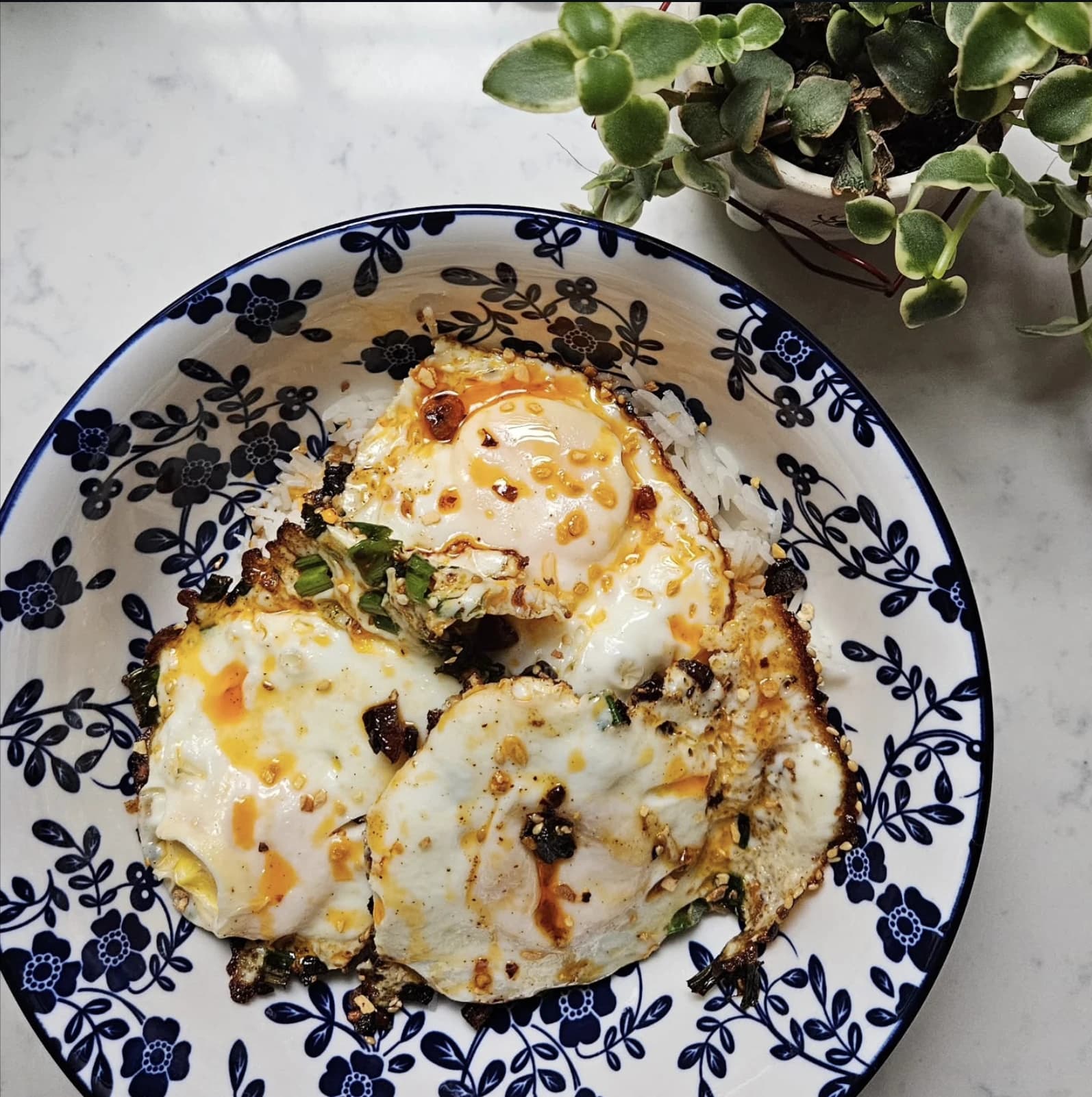 Three fried eggs over steamed rice in a blue floral bowl, drizzled with Ali's Heat Crunch Delight chilli oil.